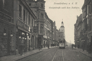 Ein altes Schwarz-Weiß-Foto einer Stadtstraße in Brandenburg, Deutschland, mit einer Straßenbahn auf Schienen, Menschen auf dem Gehweg, Gebäuden mit Fenstern, Schildern, Straßenlaternen und einem bewölkten Himmel.