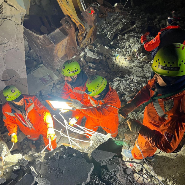 Eine Gruppe von Menschen in orangefarbenen Uniformen und grünen Helmen bei der Arbeit an Schutt von einem eingestürzten Gebäude, mit Werkzeugen und Materialien, mit einem Kran im Hintergrund.
