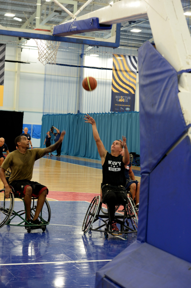 Gruppe von Menschen im Rollstuhl, die Basketball in einer Turnhalle mit einem Basketballkorb, Vorhängen an der Wand, Bannern und Deckenlampen spielen.
