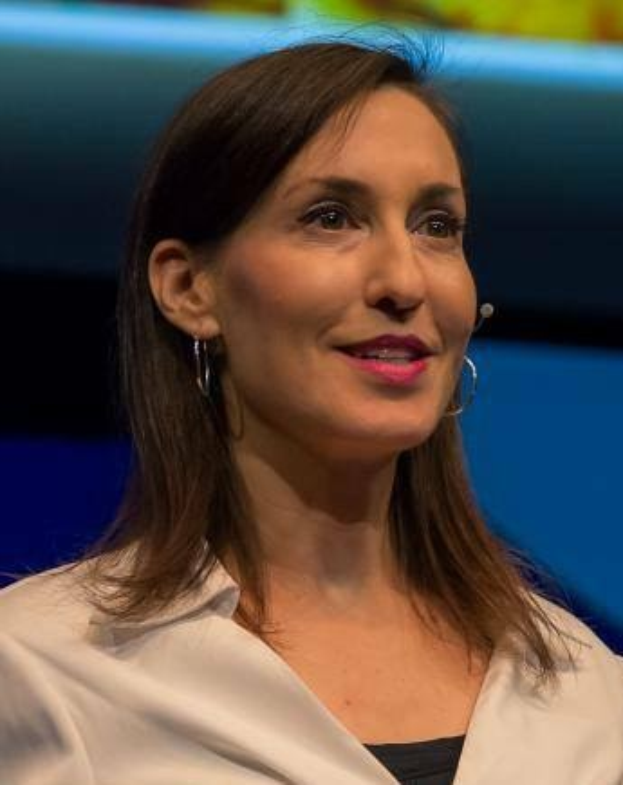 Eine Frau in einer weißen Jacke und einem schwarzen Oberteil sitzt vor einem blauen und gelben Hintergrund, trägt Ohrringe und ein Mikrofon während eines Interviews.