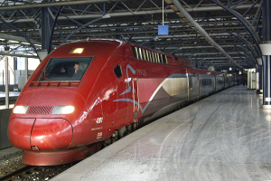 Ein roter Hochgeschwindigkeitszug steht auf einem Bahnsteig mit einigen Menschen und verschiedenen Objekten im Hintergrund.