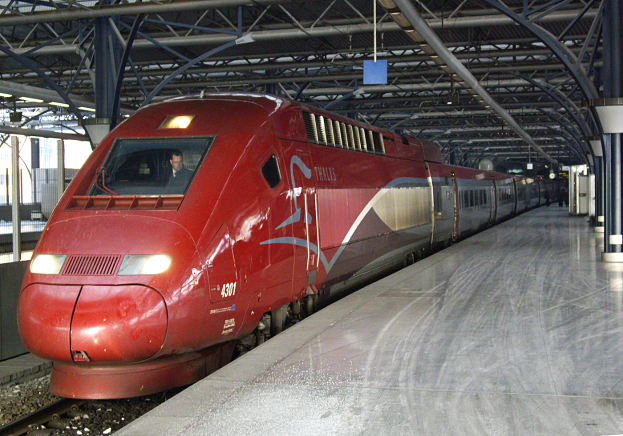Ein roter Hochgeschwindigkeitszug steht auf einem Bahnsteig mit einigen Menschen und verschiedenen Objekten im Hintergrund.