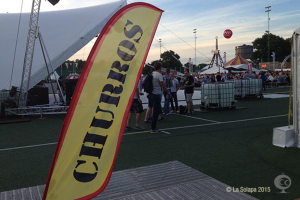 Eine bunte Faschingszeltlandschaft mit Zelten, einem zentralen Mast, Menschen, Bäumen, Strommasten, einem Himmel, Straßenbahnen auf der Straße und einem Eisengestell auf der linken Seite.