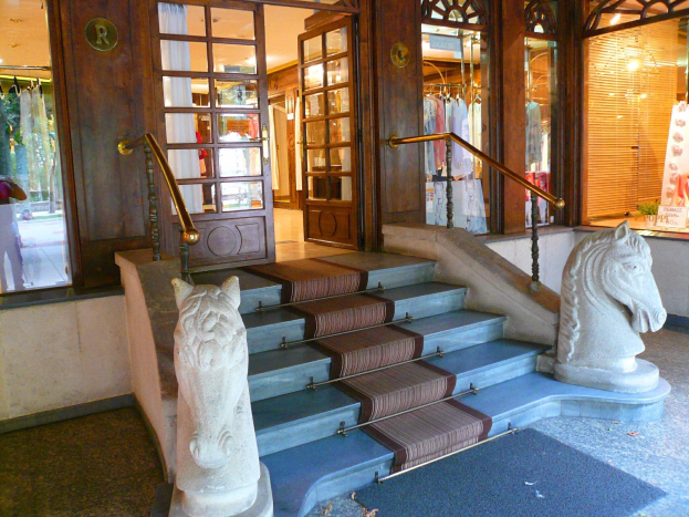 Der Eingang zu einem Laden mit zwei Pferdestatuen auf beiden Seiten der Treppe, einem Teppichboden, Glastüren, Schaufensterpuppen in Kleidern und dekorativer Beleuchtung.