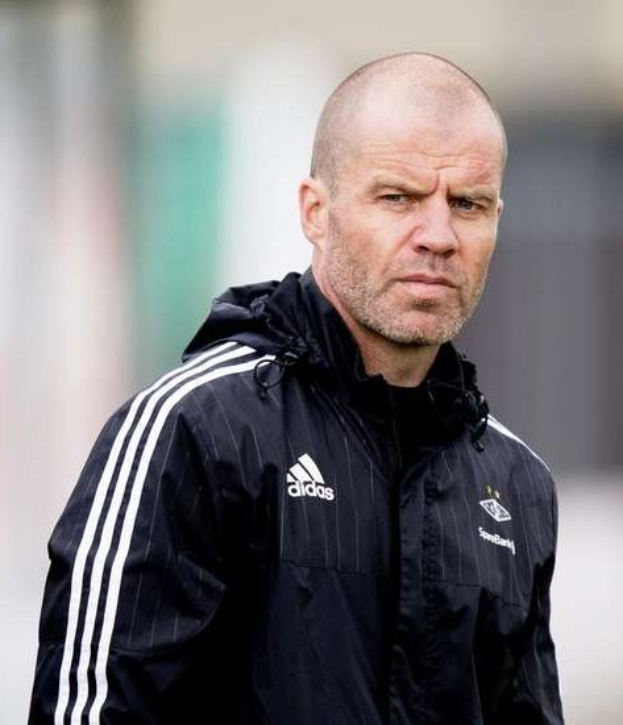 Ein kahlköpfiger Mann in einer schwarzen Jacke, der wie ein Trainer aussieht, steht auf einem Fußballfeld und trägt eine marineblaue Jacke mit weißen Streifen und einem weißen Kragen, die Hände sind verschränkt und der Gesichtsausdruck ernst.