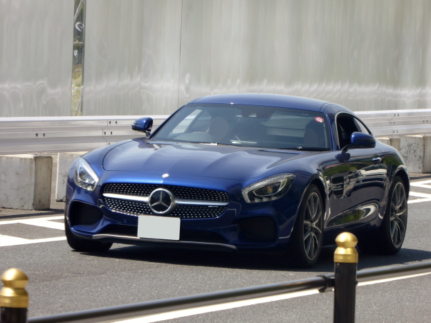 Blauer Mercedes-Benz AMG GT Coupé auf einer Straße fahrend, mit einer Wand im Hintergrund, das eine elegante aerodynamische Gestaltung, eckige Scheinwerfer, ein großes Gitter, glänzende große Räder und ein luxuriöses Lederinnenraum aufweist.