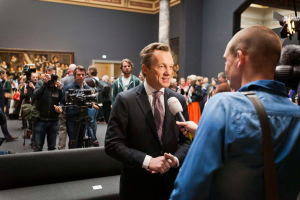 Ein Mann im Anzug und Krawatte spricht mit einem Reporter vor einer Menge, einige halten Kameras, mit einem Sofa im Vordergrund und einer Wand mit einem Foto, Lampen und einer Säule im Hintergrund, hält ein Mikrofon.