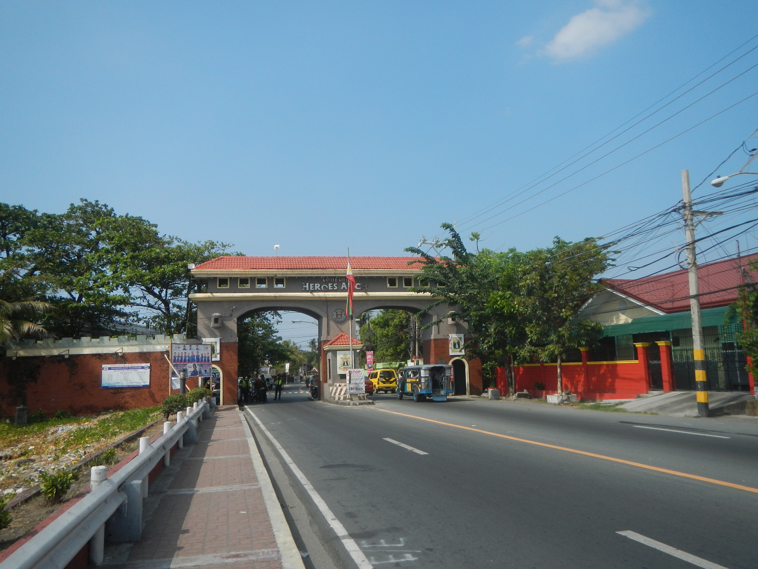 Eine zweispurige Straße mit fahrenden Fahrzeugen, ein Fußweg mit Geländern auf der linken Seite, Bäume und Gebäude auf beiden Seiten, ein Tor mit einem Namensschild, Stromleitungen an Masten und ein bewölkter Himmel.