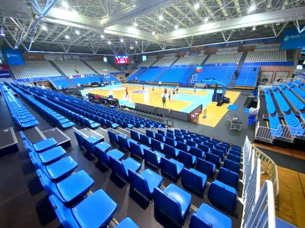 Große Hallenarena in Sarajevo, Bosnien und Herzegowina, mit blauen Stühlen, einem Basketballfeld, ein paar Menschen, Deckenleuchten, Trägern und einer Wand im Hintergrund.