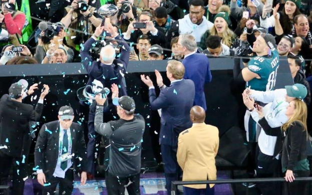 Philadelphia Eagles-Spieler feiern mit dem Vince Lombardi Trophy nach dem Sieg im Super Bowl, einige tragen Mützen und halten den Pokal, während andere Konfetti werfen, mit einem Geländer und Fotografen im Hintergrund.
