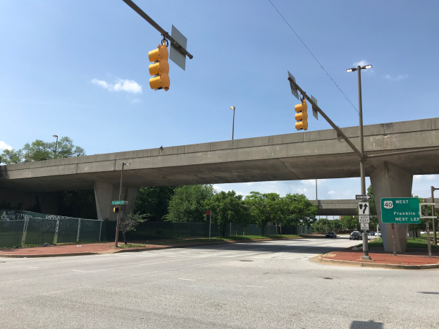 Verkehrsampel an einer Straßenrandbrücke mit darunterfahrenden Fahrzeugen, umgeben von Laternenmasten, Texttafeln, Maschendrahtzaun, Bäumen und einem bewölkten Himmel.