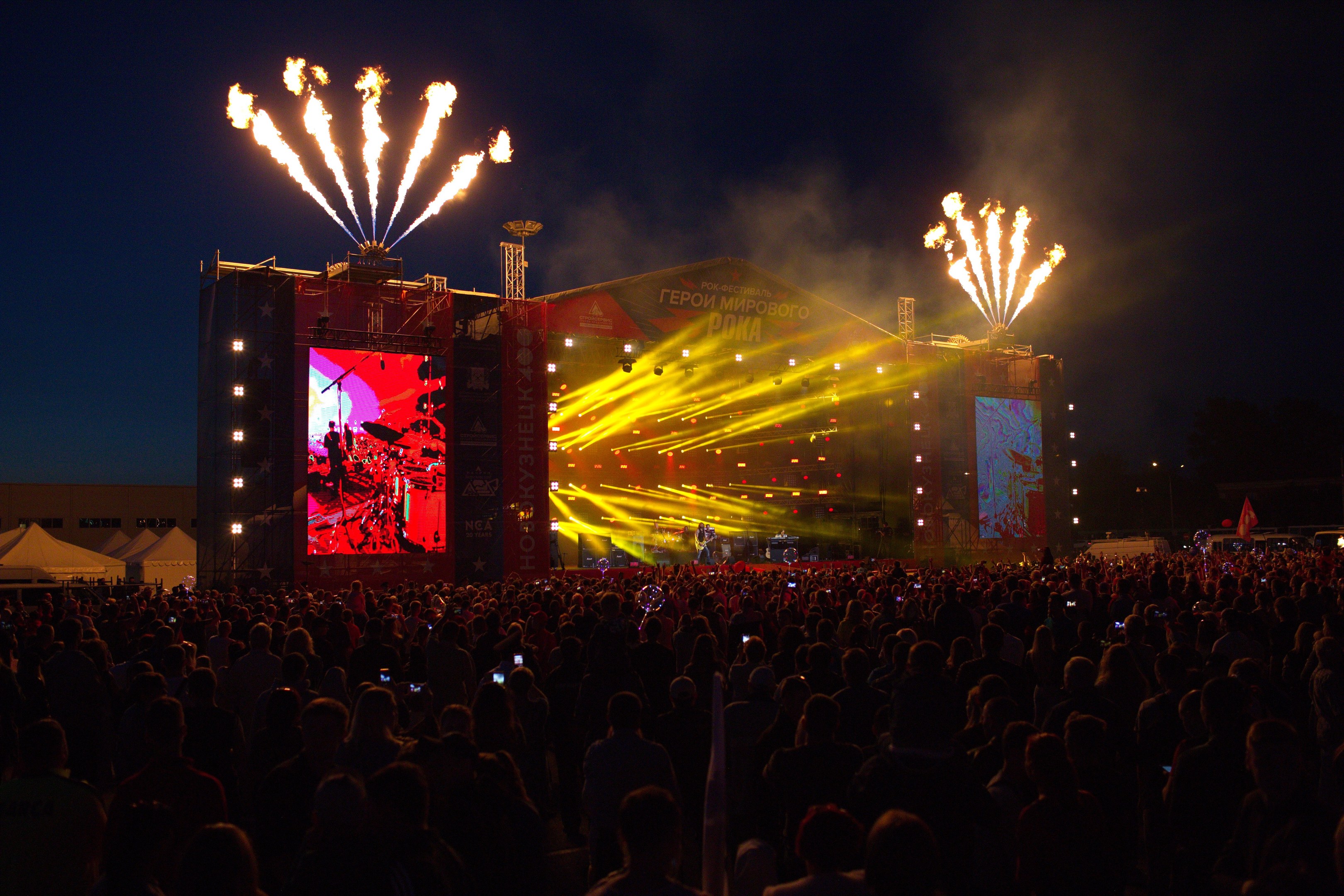 Eine große Menge bei einem Musikfestival schaut auf eine hell erleuchtete Bühne mit Zelten links und Feuerwerk am dunklen Himmel dahinter.