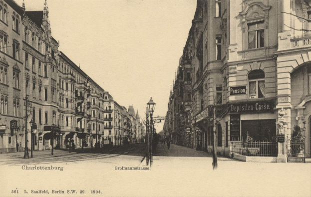Schwarzes und weißes Foto von Charlottenburg, Deutschland, das eine Stadtstraße mit Gebäuden, Straßenmasten, Lampen, Namensschildern, Kraftfahrzeugen, Fußgängern, Gittern und einem klaren Himmel zeigt.