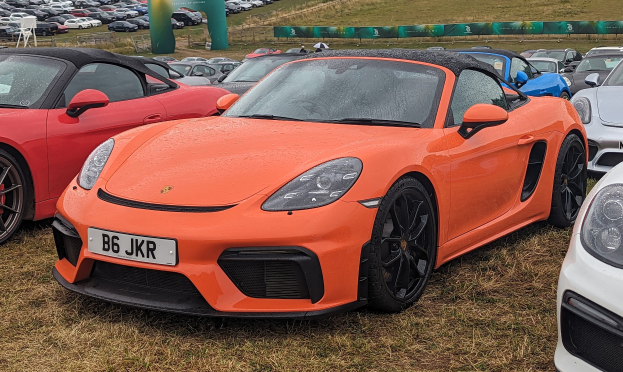 Roter Porsche 718 Boxster GTS auf dem Goodwood Festival of Speed, mit anderen Autos, einem Banner, Bäumen und einem klaren blauen Himmel im Hintergrund.