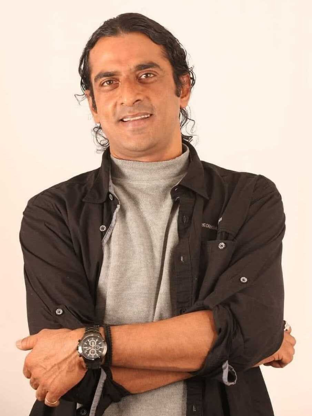 Ein Mann in einem schwarzen und aschfarbenen Kleid mit überschlagenen Armen und einer Uhr steht vor einem wei├čen Hintergrund und sieht nachdenklich aus.