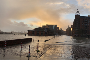 Eine ├╝berflutete Stra├če in Hamburg, Deutschland, mit Wasser auf der Stra├če, Masten, Schildern, Geb├Ąuden mit Fenstern, einer Br├╝cke und einem bew├Âlktem Himmel im Hintergrund.