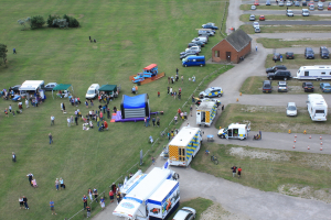 Eine Vogelperspektive eines Parkplatzes mit vielen Fahrzeugen, einigen Menschen in der Nähe von Zelten und einer zentralen Hütte sowie ein paar Bäumen auf der linken Seite.
