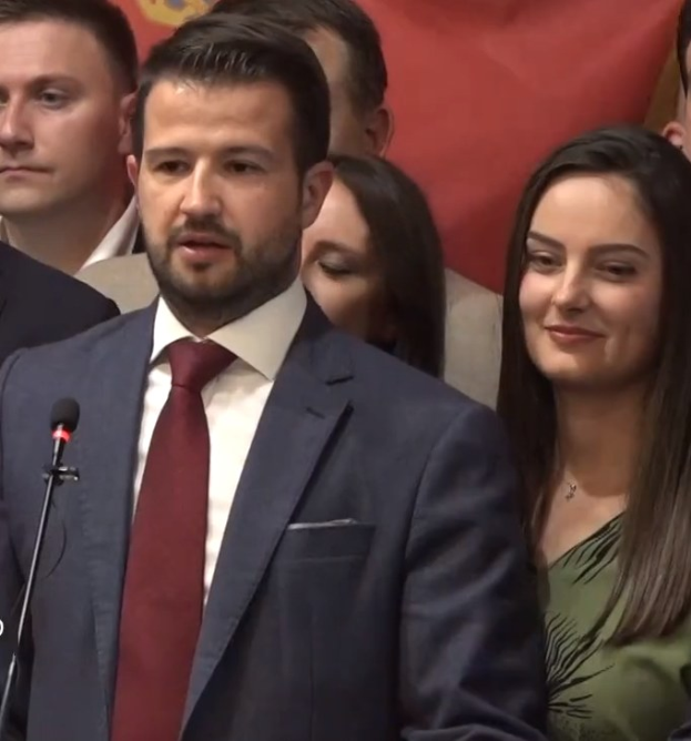 Ein Mann im Anzug mit Mikrofon spricht vor einer Menge, im Hintergrund ist eine Fahne zu sehen.