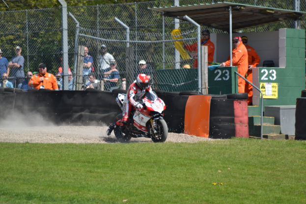 Eine Person in Helm und Handschuhen fährt ein Motorrad auf einem grasbewachsenen Feld, mit Zuschauern in Mützen und Schutzbrillen, einem Zaun, Treppen mit Geländern und einem nummerierten Schuppen im Hintergrund.
