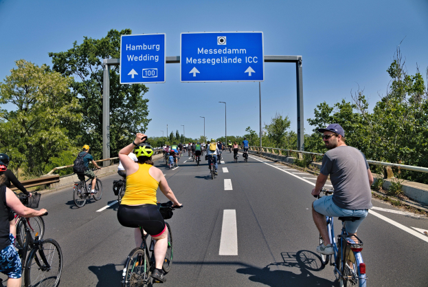Gruppe von Radfahrern mit Helmen auf einer Straße mit einer Begrenzung auf der linken Seite, Bäumen auf der rechten Seite, Laternenpfählen und einem klaren blauen Himmel im Hintergrund, mit einem Schild oben, das eine Radtour in Hamburg anzeigt.
