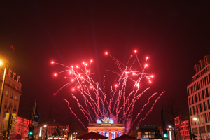 Eine Straßenansicht in Berlin am Silvesterabend, gefüllt mit Gebäuden, Bäumen, Laternen, Verkehrszeichen, Zeltplanen und Menschen, mit einem Feuerwerk-illuminierten Himmel im Hintergrund.