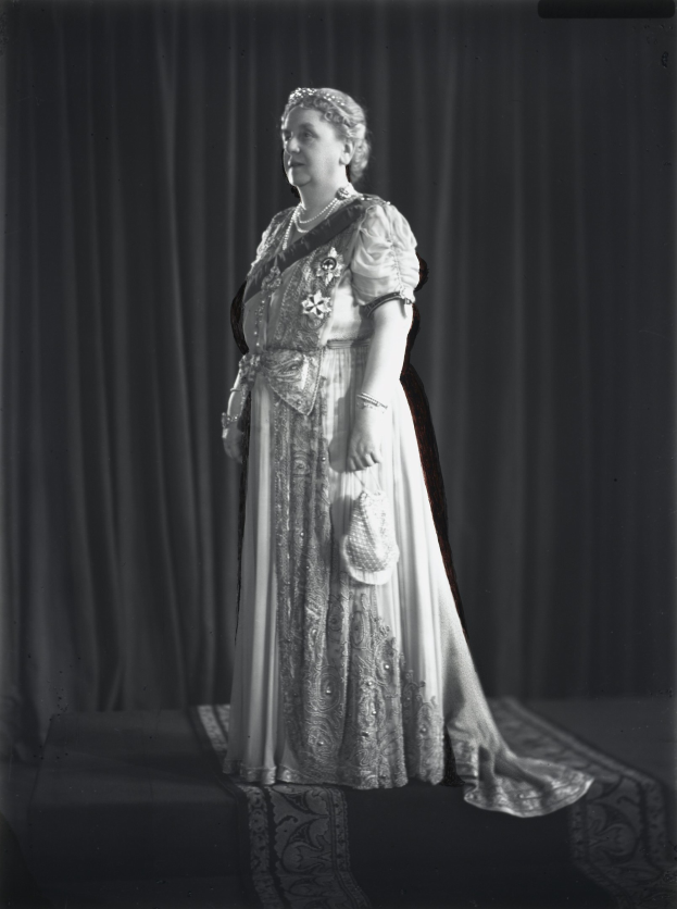 Schwarz-weiß-Foto von Queen Elizabeth II auf einer Bühne mit einem Vorhanghintergrund, die eine detaillierte königliche Robe mit einem langen Rock trägt, frisiertes Haar in einem Dutt und einen sanften Ausdruck, der zur Seite schaut.