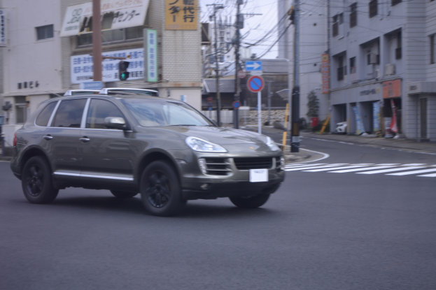 Ein Porsche Cayenne fährt auf einer von hohen Gebäuden gesäumten Stadtstraße, mit Strommasten, Schildern und Bannern, und der Himmel ist im Hintergrund zu sehen.
