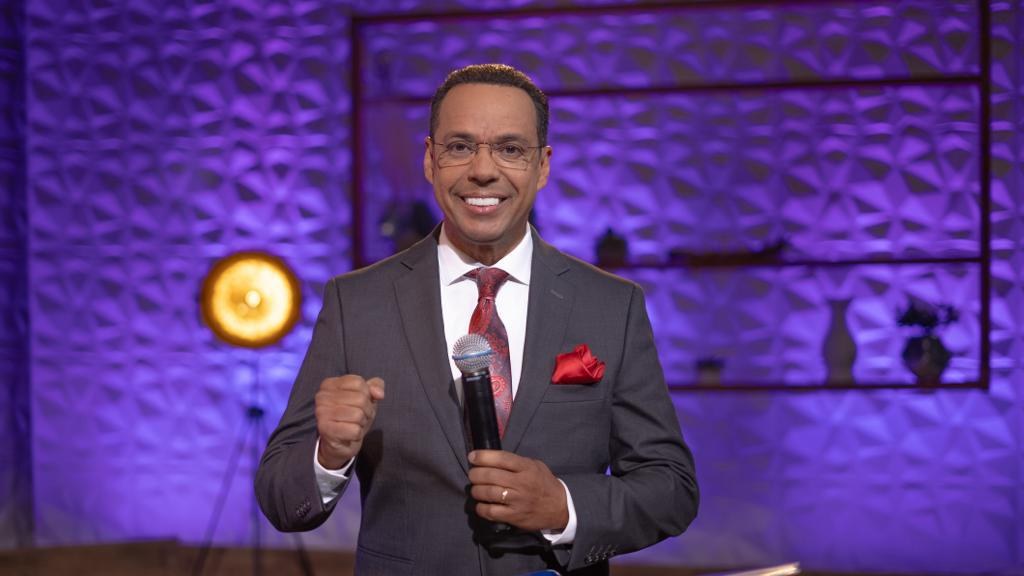 Stephen Colbert, Moderator von 'The Daily Show with Trevor Noah', steht auf einer Bühne und hält ein Mikrofon in der Hand, trägt einen Anzug, Krawatte und Brille, mit einem Ständer und Licht hinter ihm.