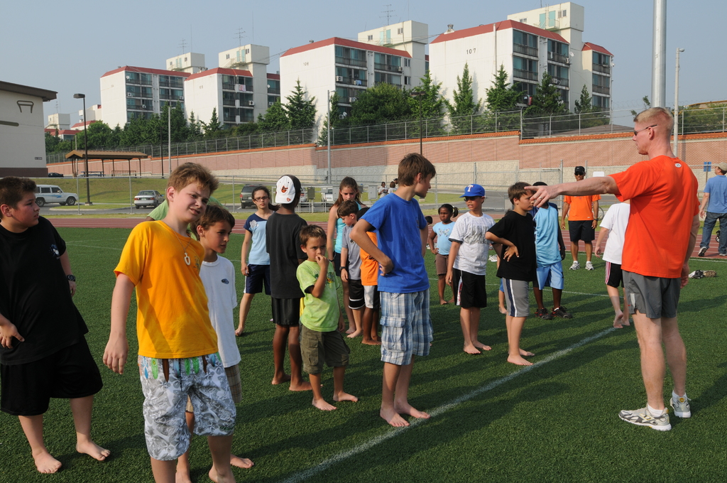 Eine grüne Fläche mit Menschen, Gebäuden, Laternen, Bäumen, Fahrzeugen und einem Zaun im Hintergrund.