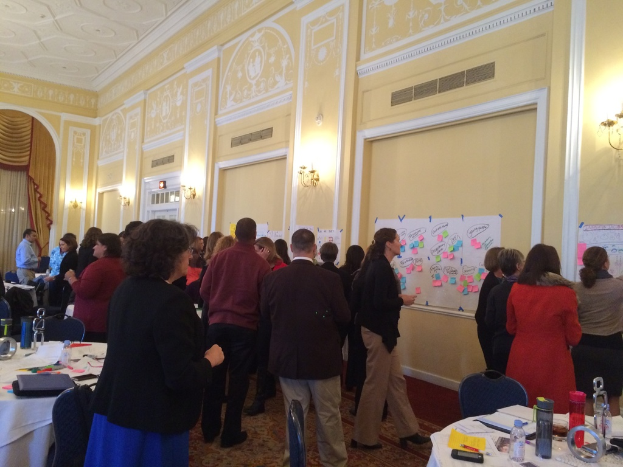 Eine Gruppe von Menschen untersucht Plakate und Charts, die an einer Wand in einem Raum angebracht sind, der wie ein Kunstmuseum aussieht.