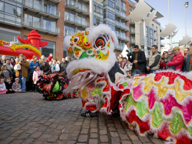 Ein lebendiges chinesisches Neujahrsfest in Amsterdam mit einem Drachentanz im Vordergrund und einer Menge Menschen drumherum, einige halten Kameras, vor einem Hintergrund aus Gebäuden, Laternenmästen und einem klaren blauen Himmel.