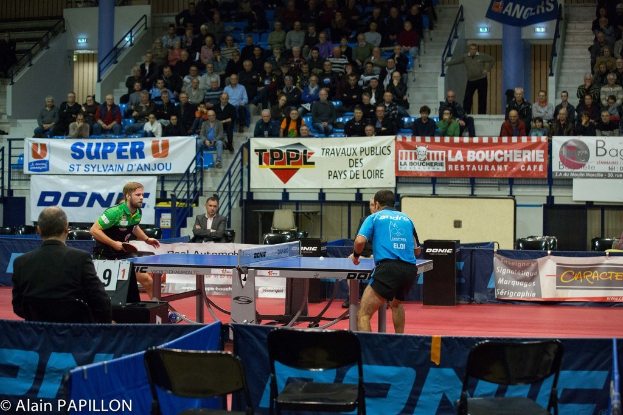 Indoor-Tischtennis-Match mit zwei Spielern, mehreren Zuschauern um den Tisch herum, leeren Stühlen und Werbeplakaten im Hintergrund.
