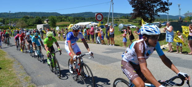 Eine Gruppe von Radfahrern fährt eine Straße entlang, während Zuschauer am Straßenrand zusehen, einige tragen Helme und Schutzbrillen, andere halten Fahnen, im Hintergrund sind Bäume, Fahrzeuge, Zelte und Gebäude unter einem klaren blauen Himmel zu sehen.