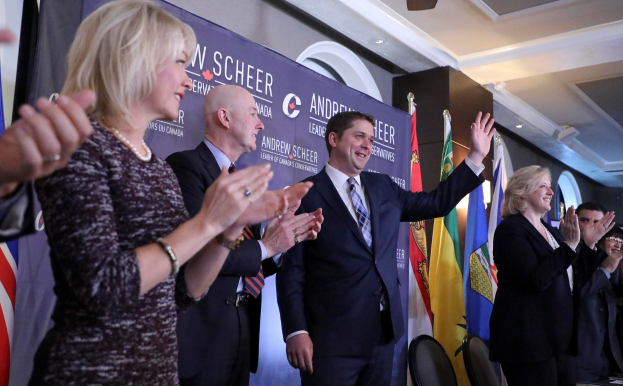 Eine Gruppe von Menschen klatscht feierlich vor Flaggen, mit einem Tisch voller Gegenstände rechts daneben, Stühlen, einem Banner und einer Wand im Hintergrund sowie Lampen und einem Ventilator auf dem Dach.