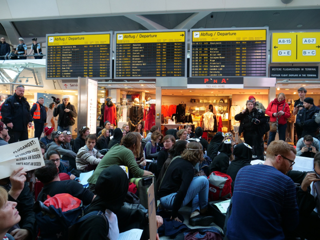 Große Gruppe von Menschen in einem Flughafen, einige sitzen mit Taschen und Papieren, andere stehen, mit Texttafeln, Kleiderpuppen und Deckenbeleuchtung im Hintergrund, was auf eine Demonstration hinweist.