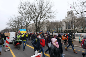 Eine große Gruppe von Menschen nimmt an einer Protestdemo auf einer Straße in Washington, D.C. teil, einige halten Schilder und Plakate, andere fahren Fahrräder, und es gibt Schilder, Bäume und einen klaren blauen Himmel im Hintergrund.