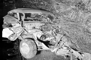 Ein Schwarz-Weiß-Foto eines schwer beschädigten Autos am Straßenrand mit einer zerbeulten Front, zersplitterten Fenstern, einem Baum im Hintergrund und verstreuten Gegenständen um die Unfallstelle.