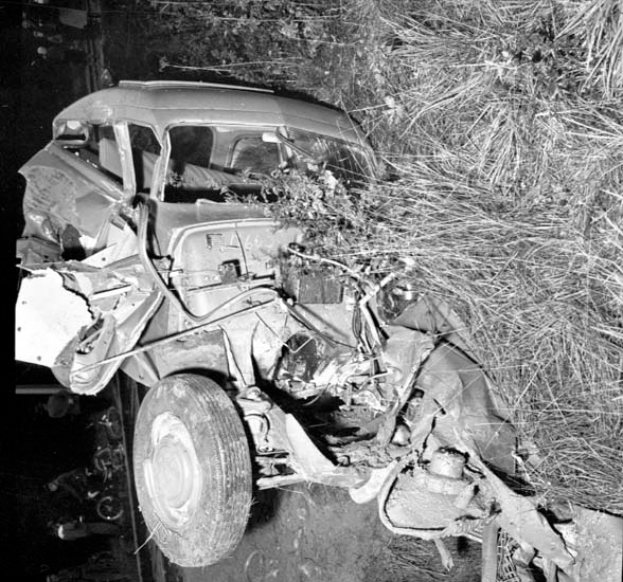 Ein Schwarz-Weiß-Foto eines schwer beschädigten Autos am Straßenrand mit einer zerbeulten Front, zersplitterten Fenstern, einem Baum im Hintergrund und verstreuten Gegenständen um die Unfallstelle.