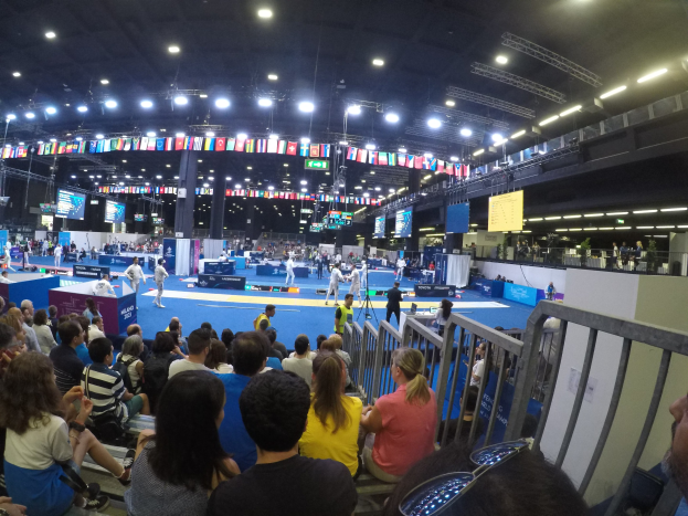 Eine große Menschenmenge sitzt und steht in einer Indoor-Sportarena mit Bildschirmen, Fahnen und Deckenbeleuchtung im Hintergrund.