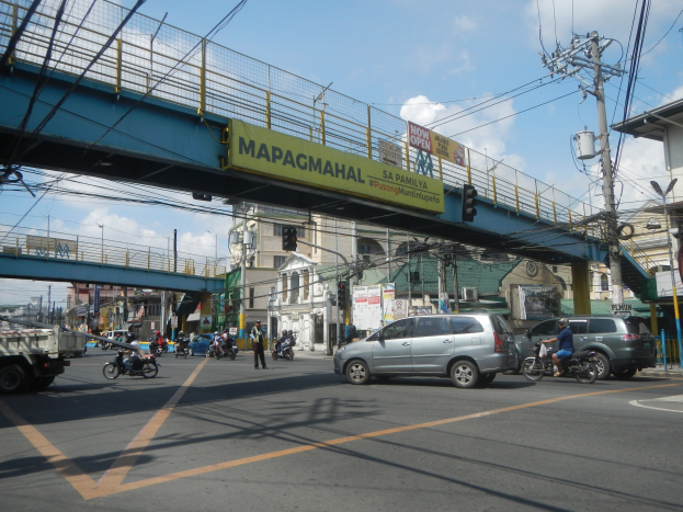 Belebte Stadtstraße mit Fahrzeugen, Fußgängern, einer Brücke mit Geländern und einem Namensschild, Strommasten mit Drähten, Laternen, Verkehrsampeln, Gebäuden mit Fenstern und einem bewölkten Himmel.