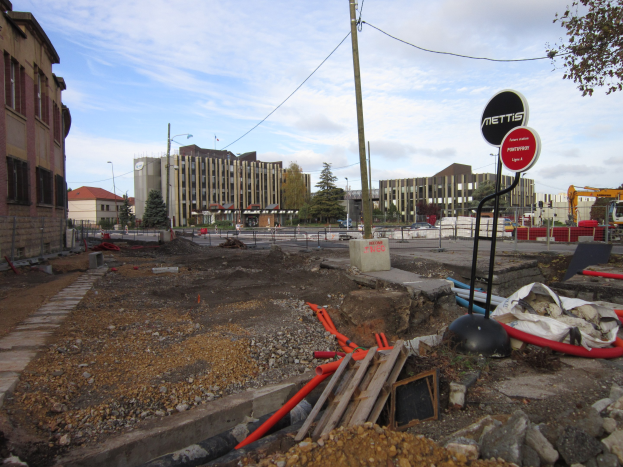 Baustelle mit verstreuten Materialien wie Steinen und Rohren, Hinweisschilder an Pfosten, ein Zaun, Fahrzeuge, Bäume, Gebäude mit Fenstern, Laternen und bewölkter Himmel im Hintergrund.
