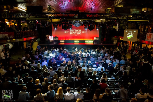 Eine große Menschenmenge sitzt auf Stühlen vor einer Bühne mit Bildschirmen und Plakaten, die von Scheinwerfern beleuchtet wird und von Geländern umgeben ist und gespannt auf den Beginn eines Online-Marketing-Rockstars-Events wartet.