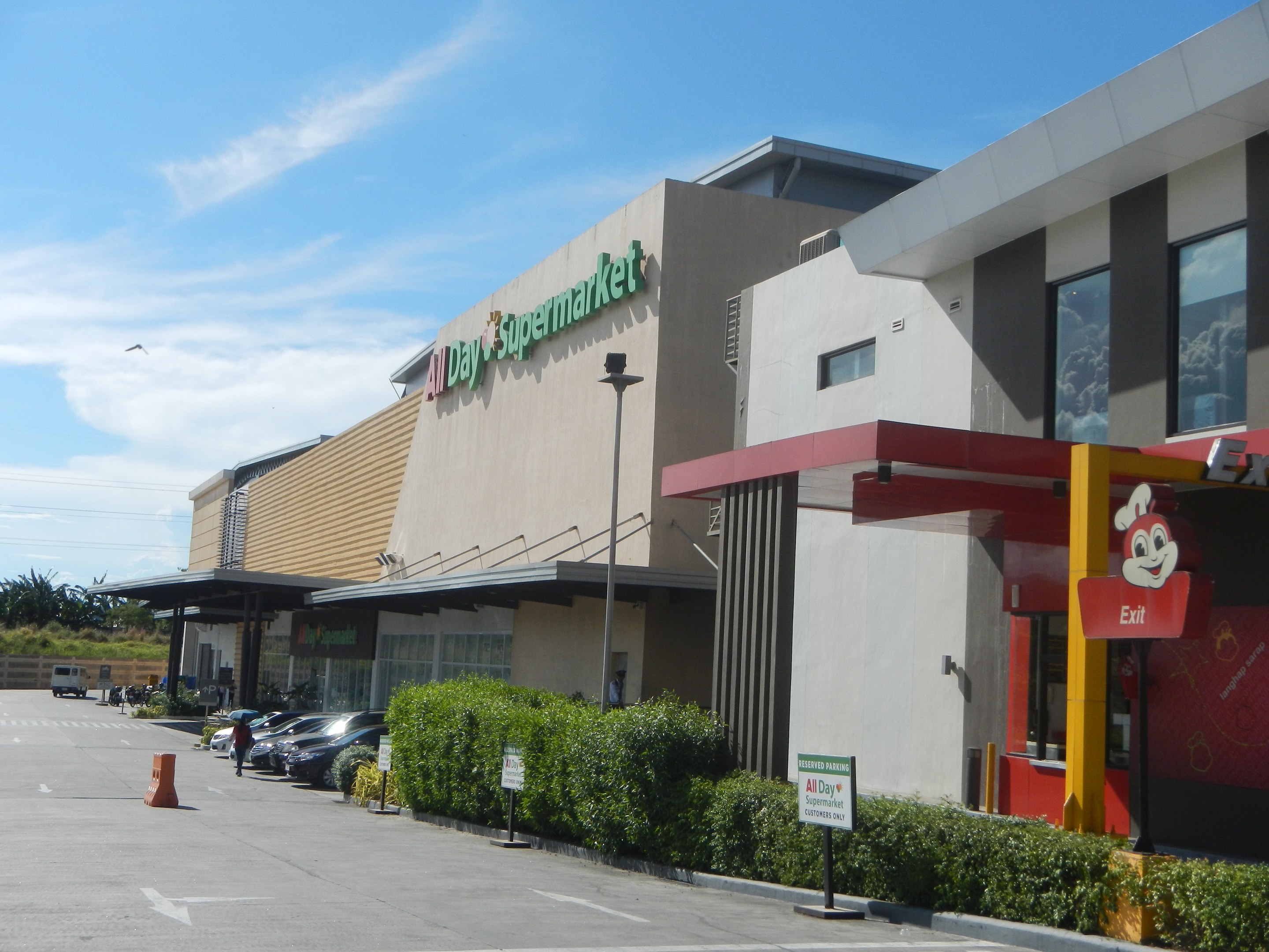 Außenansicht eines Supermarkts mit Parkplatz, umgeben von Gebäuden, Straßeninfrastruktur und Fahrzeugen unter einem bewölkten Himmel.