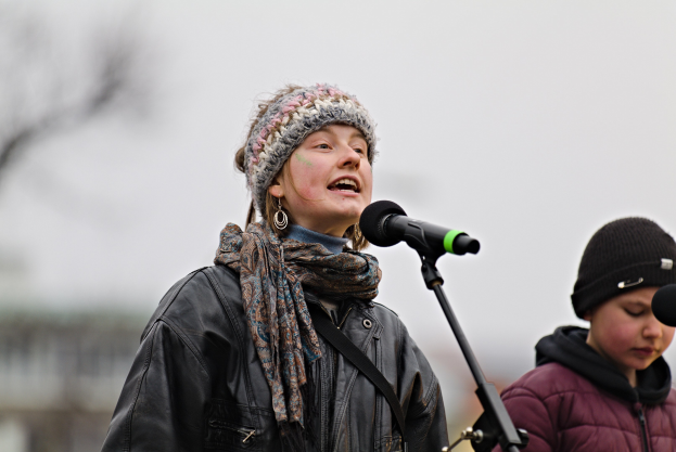 Eine Frau singt in ein Mikrofon auf der Bühne, trägt eine schwarze Jacke, einen Schal und eine Mütze, neben einer Person in einer maronenfarbenen Jacke und schwarzer Mütze vor einem unscharfen Hintergrund.