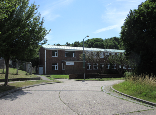 Ein Schulgebäude mit Fenstern, einer Straßenlaterne, einem Schild, einer Baumgruppe, Gras, einem Zaun und einem bewölkten Himmel, das in der Mitte eines Parks umgeben von saftigem Gras steht.