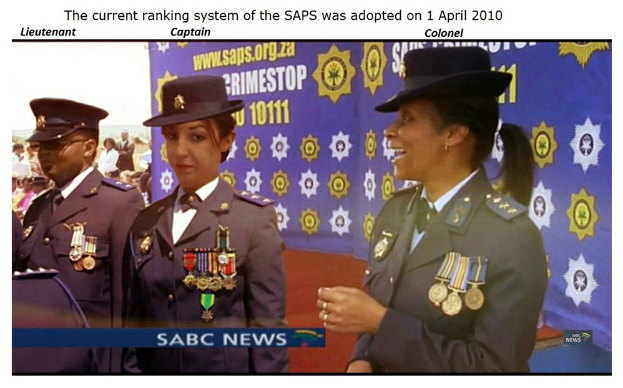 Gruppe von Menschen in Uniformen und Hüten vor einem Banner mit Text über und unter dem Banner "Das aktuelle Rangsystem der SAP wurde am 1. April 2010 übernommen".