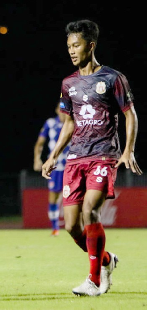 Ein Mann in einem rotbraunen Hemd und roten Shorts spielt Fußball auf einem Feld, mit einer weiteren Person im Hintergrund und einer Tafel mit Text.