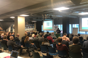 Eine große Gruppe von Menschen sitzt auf Stühlen und schaut auf eine Projektionsfläche, während ein Redner neben einem Mikrofon und einem Gerät auf einem Ständer steht, in einem Raum mit Säulen, Fenstern, Deckenleuchten und einer Tafel mit Text.