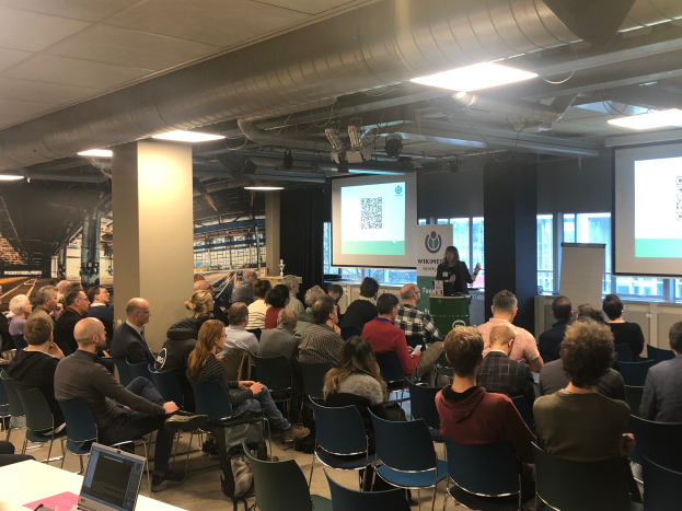 Eine große Gruppe von Menschen sitzt auf Stühlen und schaut auf eine Projektionsfläche, während ein Redner neben einem Mikrofon und einem Gerät auf einem Ständer steht, in einem Raum mit Säulen, Fenstern, Deckenleuchten und einer Tafel mit Text.