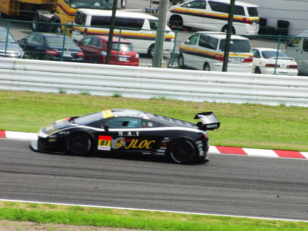 Ein schwarzer Sportwagen fährt auf einer Rennstrecke, umgeben von Gras, Zaun, Pfosten, Maschendraht und anderen Fahrzeugen, mit einer Wand im Hintergrund.